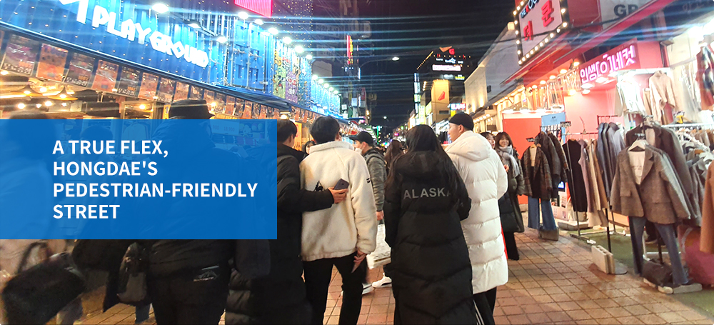 Close-up of a crowd walking up Hongdae's shopping street at night with the clothing store, Playground, on the left with the title: A true flex, Hongdae's pedestrian-friendly street