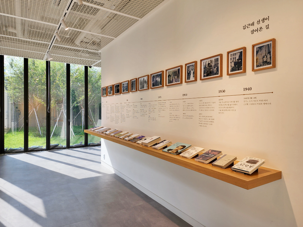 A historical rundown by year of Kim Geun-tae is written on the wall accompanied by pictures with related works displayed on a long wooden shelf.