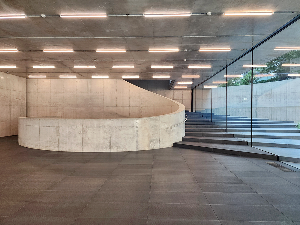 A concrete spiral wall on the left, and the first- floor lobby from where the outdoor garden can be seen through the glass window
